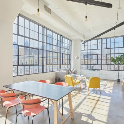Modern loft living space with dining table, orange dining chairs, yellow accent chairs, gray couch, large industrial windows, concrete floors.